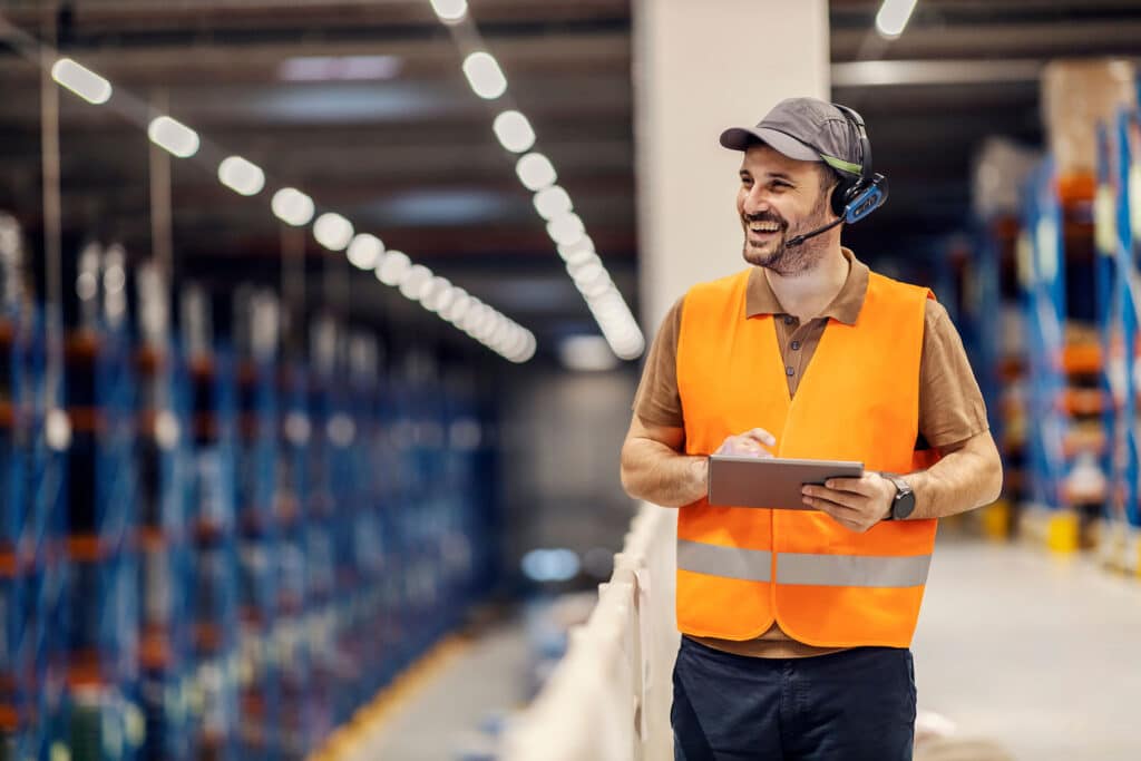 A happy logistics worker is using headset and tablet for work in storage.